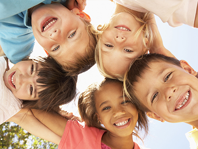 The image shows a group of six children of various ages and genders, smiling at the camera, with their arms around each other's backs, set against a bright sky background.