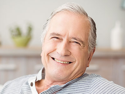 The image shows an older man with white hair, smiling broadly, wearing a blue shirt, seated comfortably with his arms crossed, in front of a homey background that includes a plant and a wall decoration.