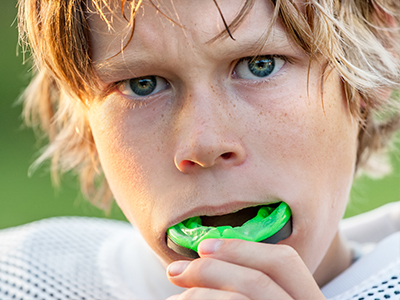 The image features a young male with blonde hair, wearing a football uniform with a mesh front, sitting on a field with his mouth open towards a green object, possibly a piece of sports equipment or a toy.