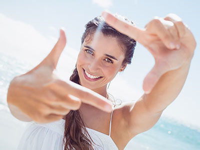 The image shows a woman taking a selfie with her finger pointing at the camera, set against a sunny beach backdrop.