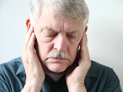 The image shows an elderly man with his eyes closed, holding his face with both hands, appearing to be deep in thought or experiencing discomfort.